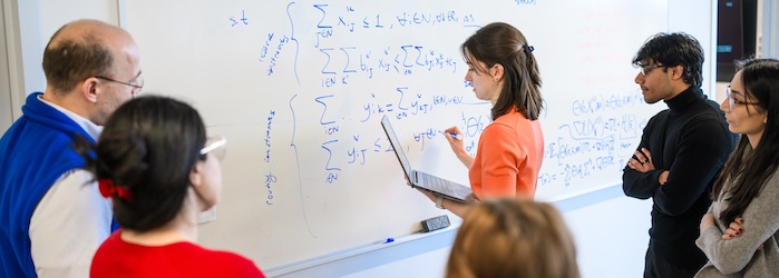 A graduate student in WPI's data science program writes on a whiteboard while other students and faculty watch
