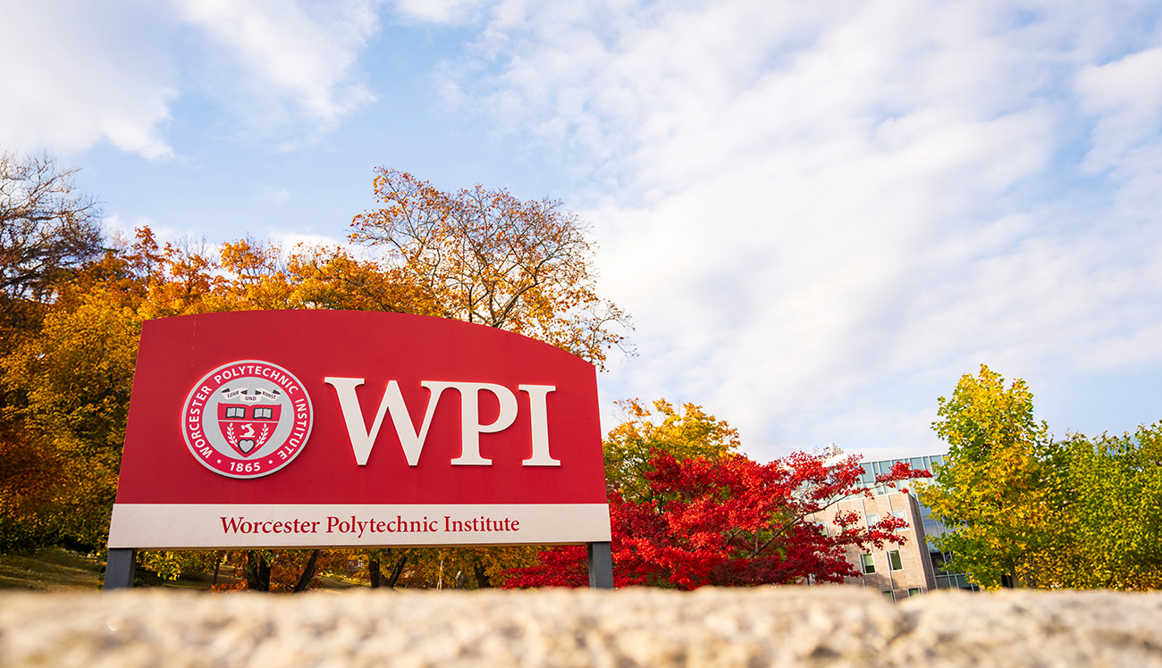 View of the entrance to WPI Campus with large red logo signage and autumn trees in the background with campus buildings in the distance.