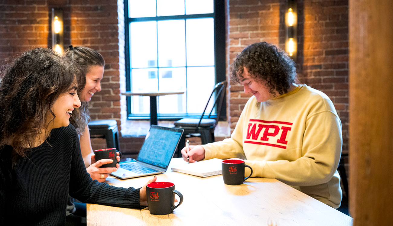 A scene of WPI students and the owner of Fuel Coffee enjoying working at a table in the coffee shop.