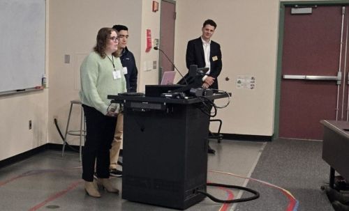 Two people stand at a lectern in a classroom while an onlooker stands nearby.