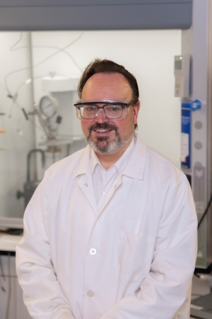 Scientist standing in front of lab equipment, wearing white lab coat.
