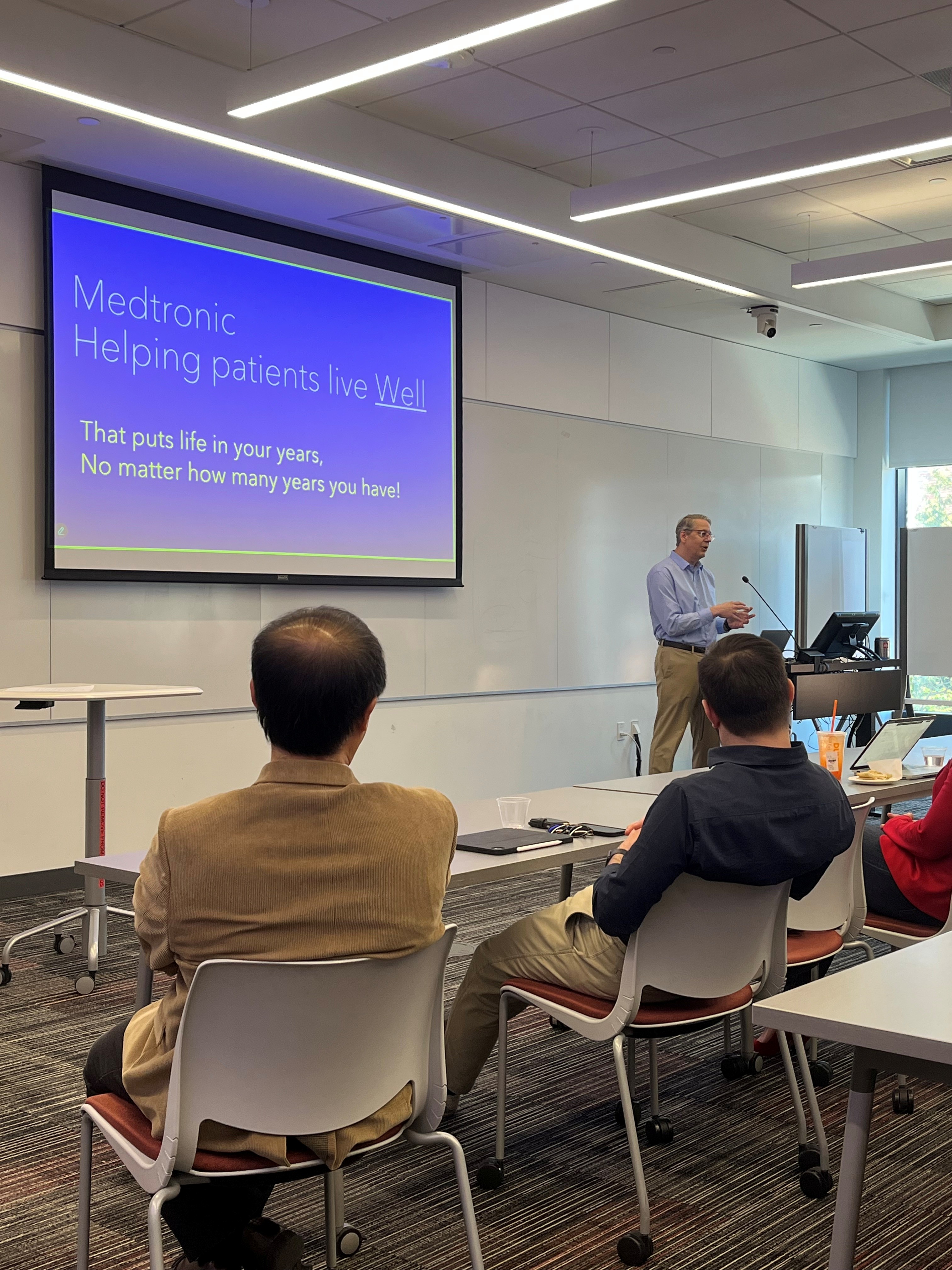 Image of Bill Peine standing at a lectern beside a projection of a blue PowerPoint slide that says "Medtronic - helping patients live well. That puts life in your years no matter how many years you have left!" Two men sit in chairs facing away from the camera towards the presentation.