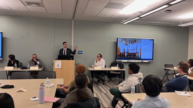 picture of the first Surgical Innovation Collaborative program information workshop, showing a male surgeon presenting from a podium to a room of students, with three other surgeons seated at tables next to him