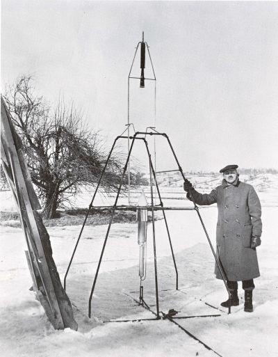 black and white photo of Robert Goddard standing outside in the snow next to a rudimentary rocket launch