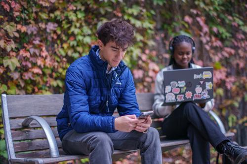 two students sitting on a bench, one looking at a cell phone, the other looking at a laptop.