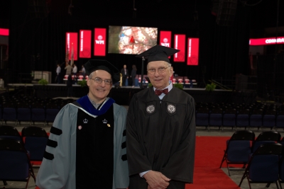 Professor and student in academic regalia standing in arena in front of graduation stage