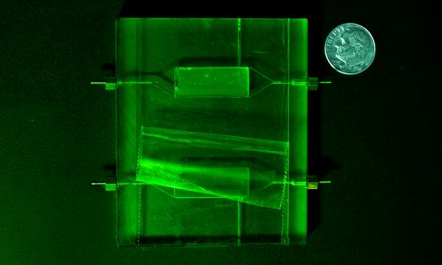 A microfluidic device appears in green light next to a dime.