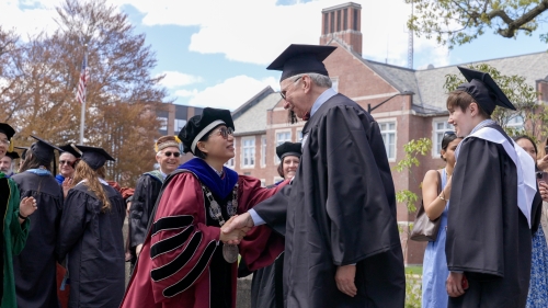 WPI President in academic regalia greeting graduating student with a handshake as other students and faculty look on