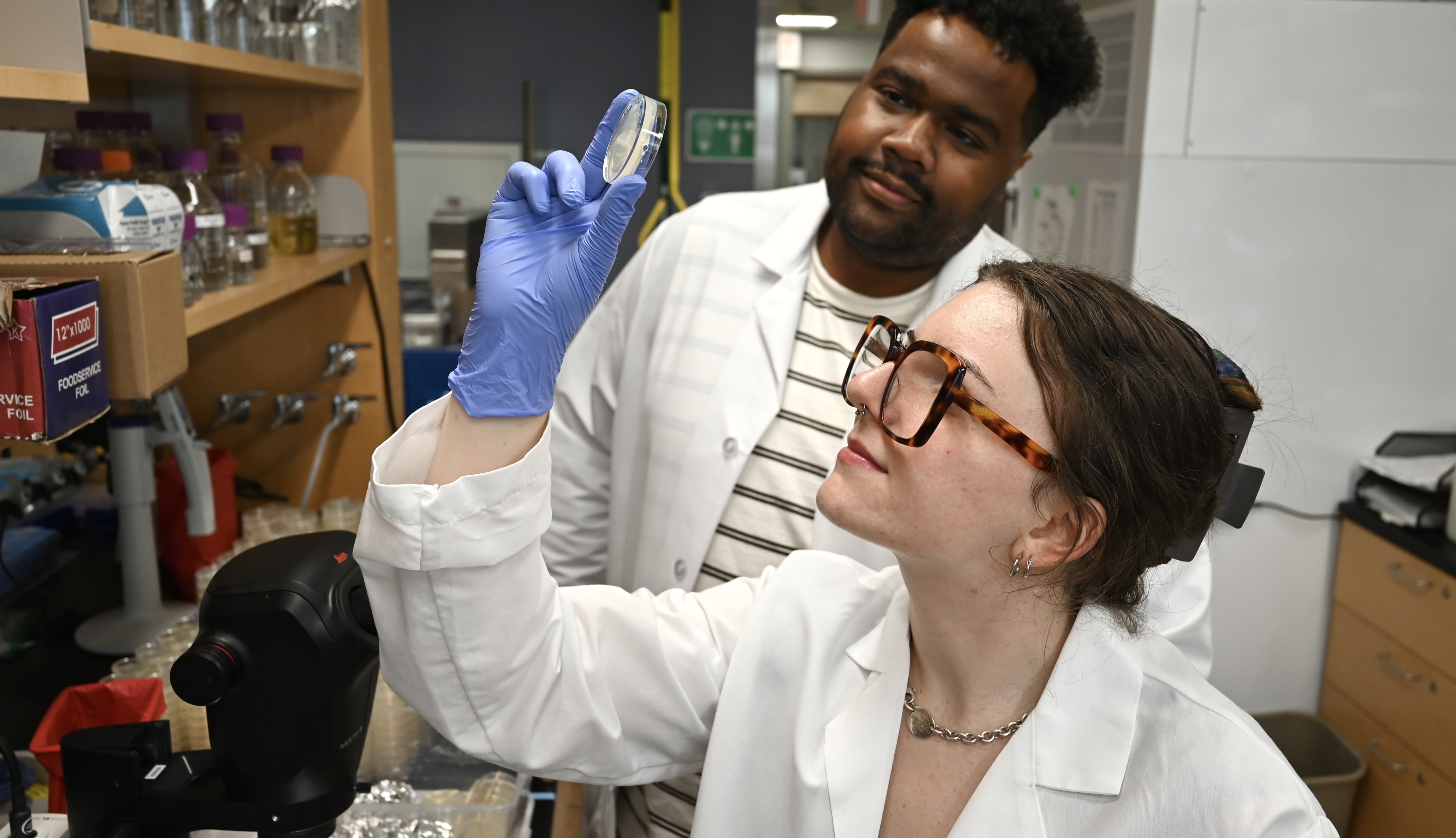 a student holds up at a petri dish and looks at the contents while a professor watches