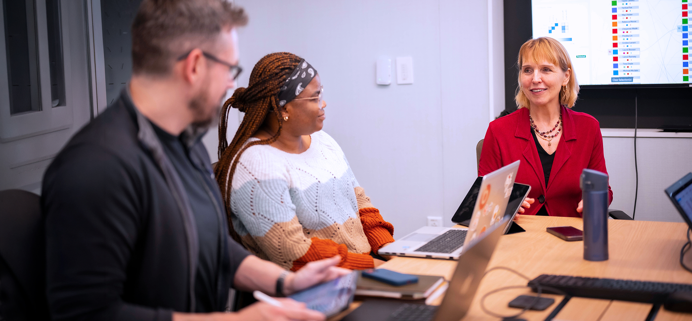 Three people participate in a meeting around a conference table with laptops and tablets, while a data visualization is displayed on a screen behind them.