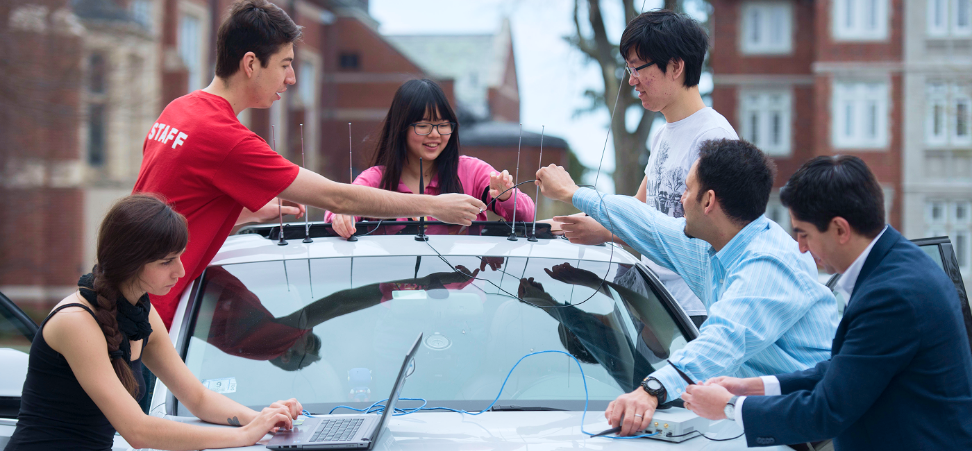 A group of students and researchers work together around a vehicle, connecting antennas and electronic equipment while using a laptop.