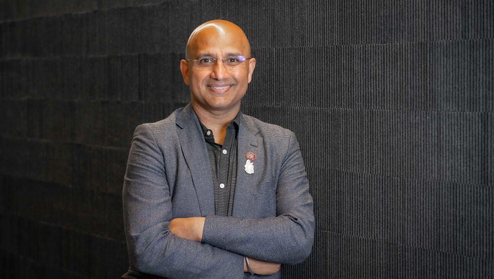 Man wearing eyeglasses and suit stands in front of a wall.