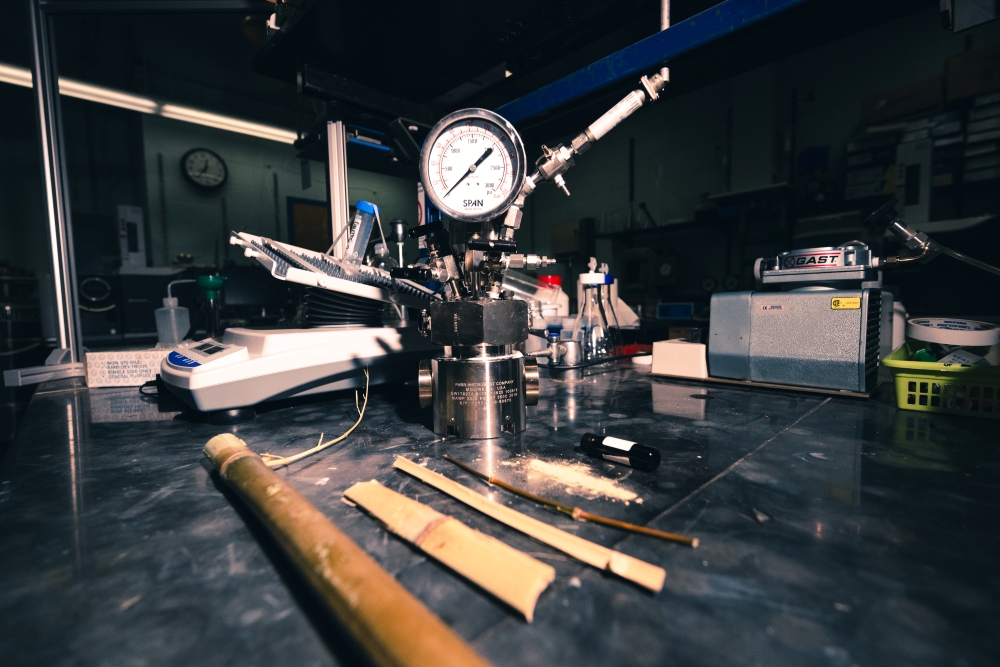 A laboratory bench with items on it including bamboo stalks, crushed bamboo powder, a vial of biocrude oil, and a metal reactor