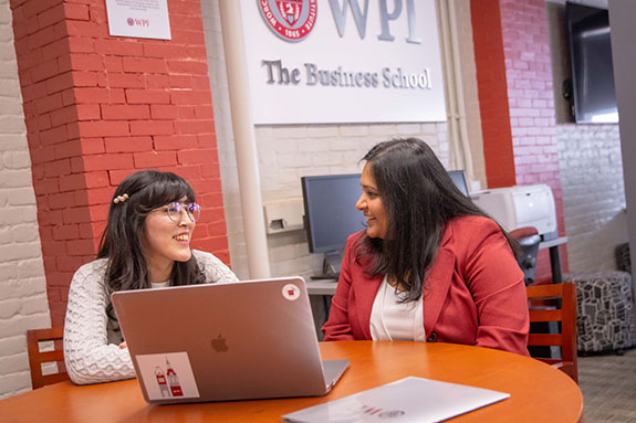 WPI Business School professor meeting with student at table in front of a red brick wall with the WPI logo in the background