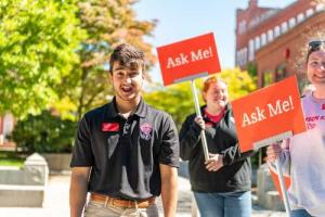 A student tour guide laughs while two others stand behind him with signs that say "Ask me."