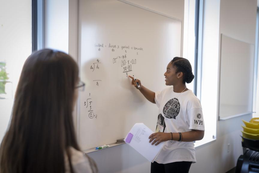 Student in a WPI T-Shirt writing a math problem on a white board explaining to another student