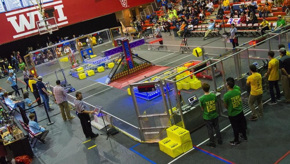 A basketball court setup for a robotics competition. Teams watching the competition and people in the stands.