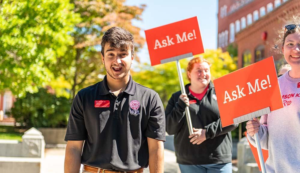 An admissions associate smiles with two red "Ask me!" signs held up in the background.