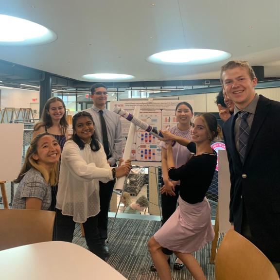 A large group of students in front of their project poster boards.  Two students in the middle are dueling using their rolled up posters as faux weapons