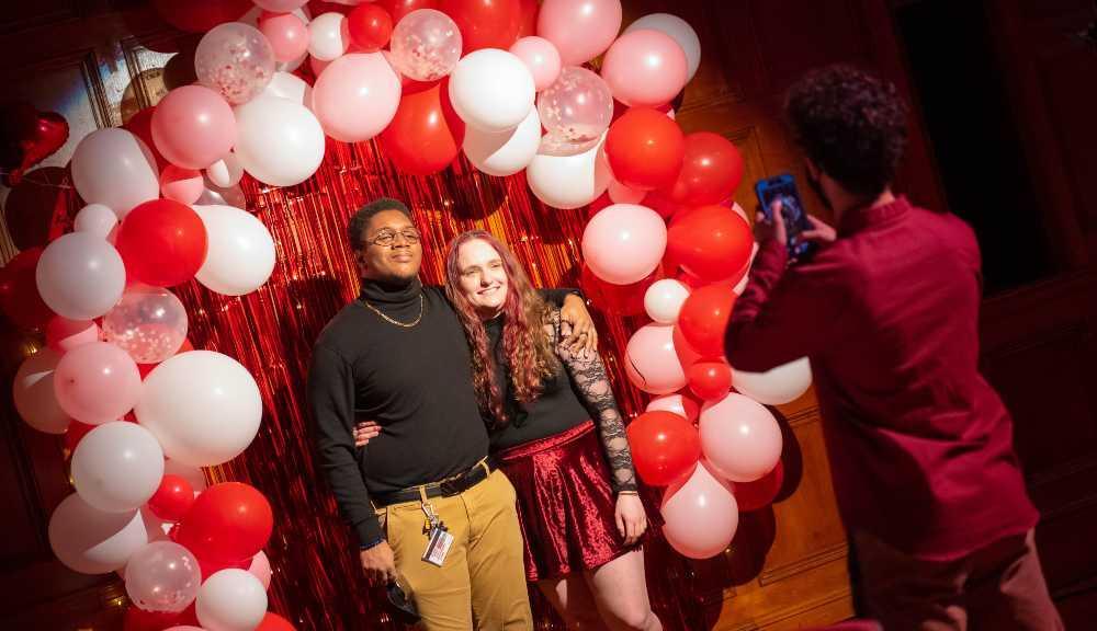 A photo of two students getting their photo taken under an arch of pink balloons