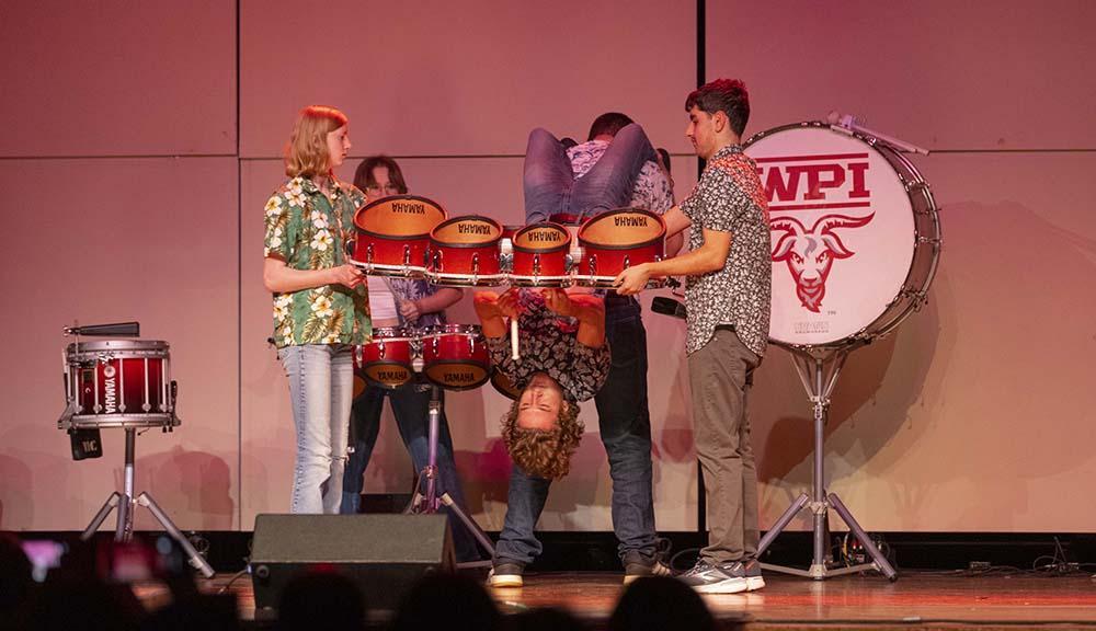 A student playing the drums while he and the drumset are being held upside down by other students