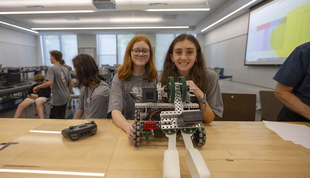 Two students smile for the camera with a small robot in front of them.