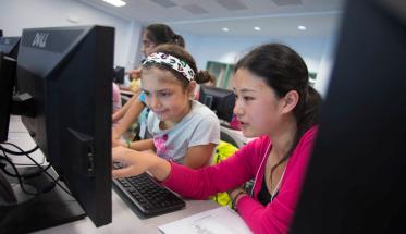 Two young girls sit next to each other and observe the content on a computer screen.