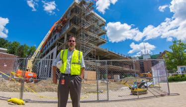 Jim Bedard wears a fluorescent yellow safety vest over a light blue button-up shirt and gray slacks. He is holding a white hard hat under one arm, and is standing in front of the Foisie Innovation Studio construction site.