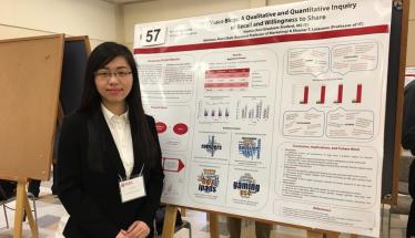 Huimin Ren is dressed in a black pantsuit and white shirt, and is standing next to a poster for her project, "Video Blogs: A Qualitative and Quantitative Inquiry of Recall and Willingness to Share."