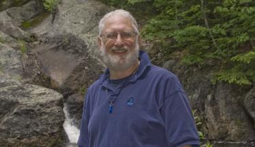 Roger Gottlieb stands in front a rock formation with running water and green foliage in the background. He's smiling, has white hair and beard, and is wearing glasses and a dark blue polo shirt.