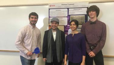 Four students (two young men, a young woman, and another young man) pose, smiling, in front of their poster on chemical treatment. The young man on the left is holding a blue shovel.