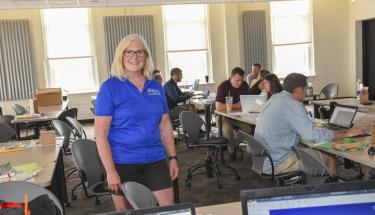Martha Cyr stands in the middle of a classroom with students and faculty members working on laptops in the background. She is smiling and has blonde hair, and is wearing glasses, a short-sleeved dark blue shirt, and black shorts.