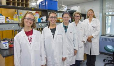 Four young women (part of the iGEM team) in front of a lab station, with Natalie Farny standing behind them. They're all smiling and wearing white lab coats and safety glasses.