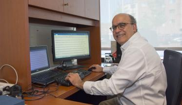 Joe Sarkis sits at his desk with two computer monitors in front of him. He's got one hand on the keyboard and one on the computer mouse, and is looking over his shoulder and smiling. He has gray hair and is wearing glasses.
