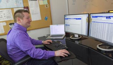 Craig Shue sits in front of two computer monitors and a laptop. He's smiling, and is wearing a purple shirt and dark blue jeans.
