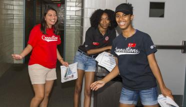 Three female students prepare for move-in day. They're all smiling, and are wearing red and blue T-shirts.