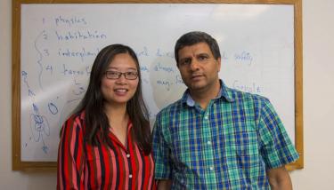 Shanshan Rodriguez and Rudra Kafle stand in front of a white board. Shanshan is wearing a pink, red, black, and brown striped blouse; Rudra is wearing a blue, green, and pale yellow checkered shirt.