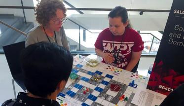 Two attendees of the Games for Change Festival gather around a table to play a board game developed by a WPI student, who looks on to provide instructions and tips.