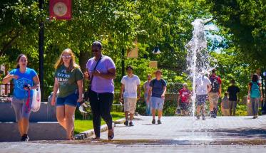Students walk past the Fountain in the center of campus on a sunny day with green foliage behind them.