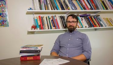 Geoffrey Pfeifer sits in his office. Two bookshelves full of books are behind him, as well as a small pile of books on the table in front of him. He's wearing glasses and a blue button-up shirt.