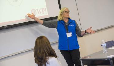 Martha Cyr stands in front of a projection and addresses a class. She's gesturing toward the projection, has blonde hair, and is wearing glasses, a blue vest, gray shirt, and black pants.