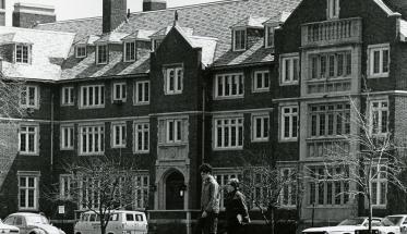 A black and white photo of Sanford Riley Hall, with two students walking in front of it. Several old cars are parked along the front of the building.