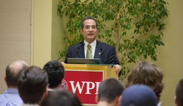 Martin Burt addresses a crowd standing at a podium emblazoned with WPI's logo. He is wearing a dark suit, white shirt, and green and gold tie.