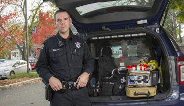 Officer Fuller in front of a WPI Police cruiser. He's wearing a dark navy police uniform, and crisis management equipment is loaded into the back of the vehicle.