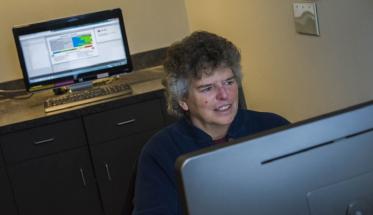 Beth Wilson studies a computer screen, with another screen behind her. She has gray hair, and is wearing a dark blue sweater and red shirt.