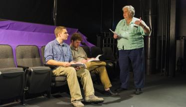 Mike Ciaraldi (right) discusses a scene in the Little Theatre with two male students who are seated in theatre chairs. They're all holding scripts; Ciaraldi is talking, one student is looking at him, and one is looking at the script.