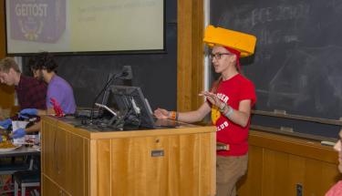 A student wearing a red shirt and yellow cheese head stands at a wooden podium, while two other students prepare cheese in the background.