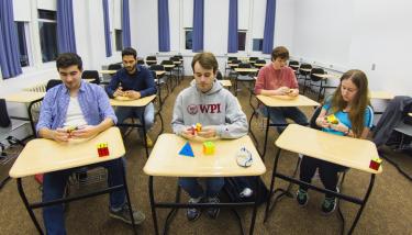 Five students sit at desks, each holding a Rubik's cube and getting ready to solve the puzzle.