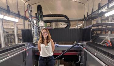 Amy Peterson stands inside of a large 3D printer. She is smiling and wearing safety glasses, a white blouse, and blue-and-white polka dot pants.
