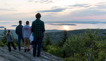 Picture of students staring into the harbor from the top of a mountain. Clouds and trees are visible in the distance.